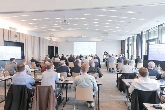 Viele Personen sitzen an Tischreifen und hören einem Vortrag zu