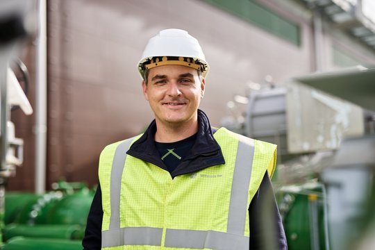 Männliche Person schaut frontal in die Kamera, trägt Arbeitskleidung, eine Warnweste und einen Helm