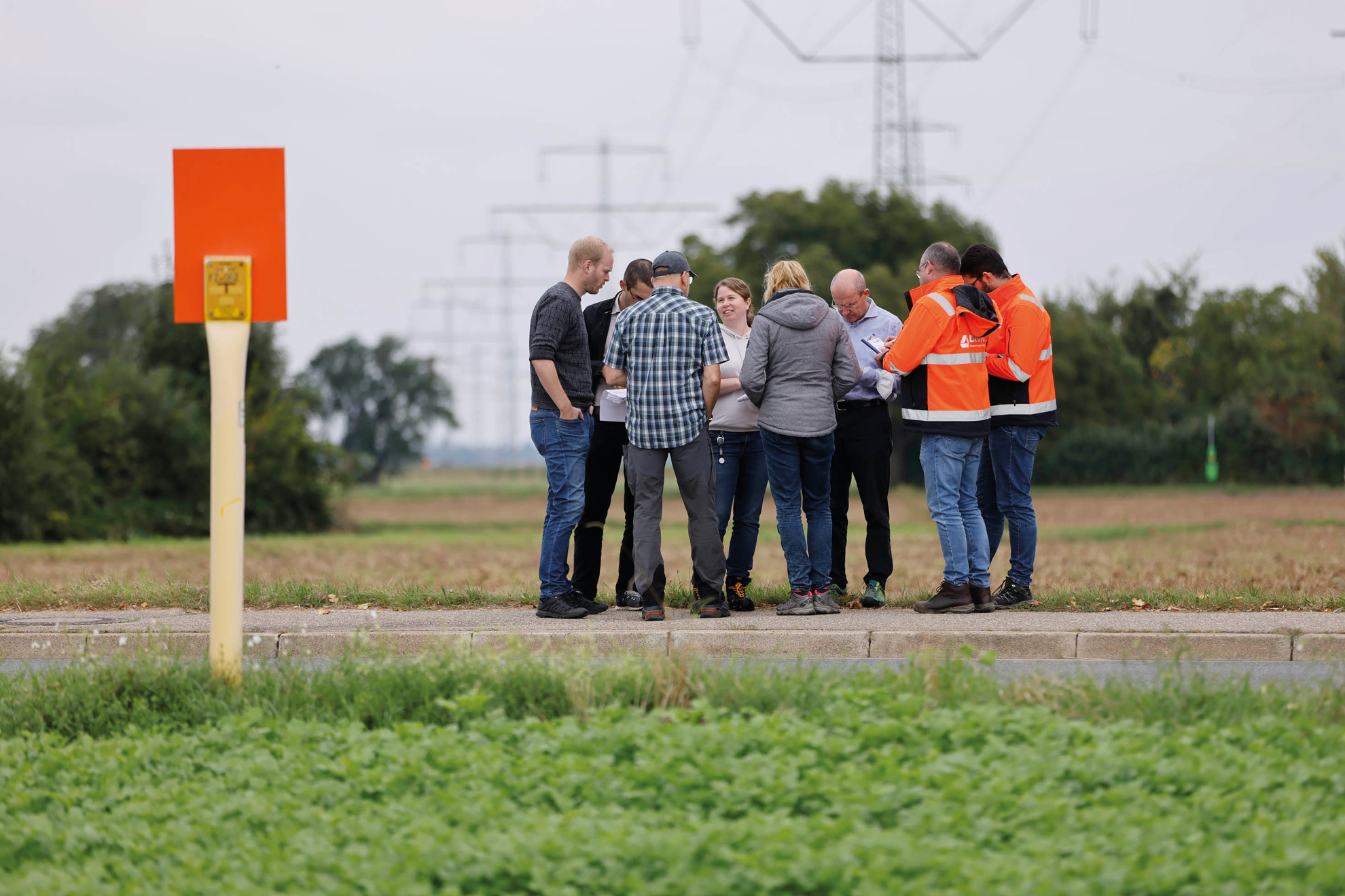 Links im Vordergrund steht eine Flugtafel, daneben ist eine Personengruppe auf einem Feldweg stehend zu sehen